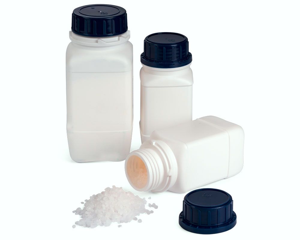 White plastic containers with dark blue screw caps. In front of it is an open container from which white coarse-grained granules fall out